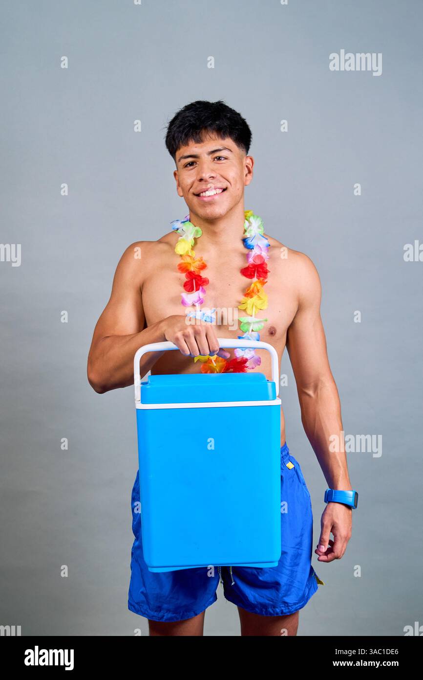 Photo studio d'un jeune homme souriant portant un maillot de bain bleu et un lei hawaïen, tenant une glacière portable sur un fond gris Banque D'Images