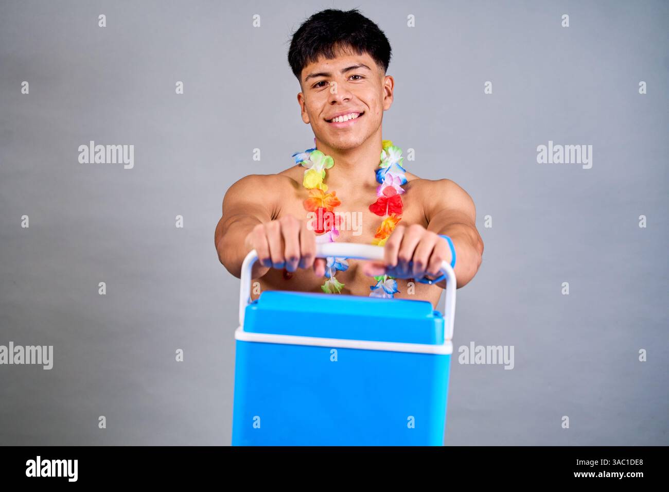 Photo studio d'un jeune homme musclé portant un lei hawaïen tenant une glacière portable, souriant et se préparant pour les vacances d'été Banque D'Images