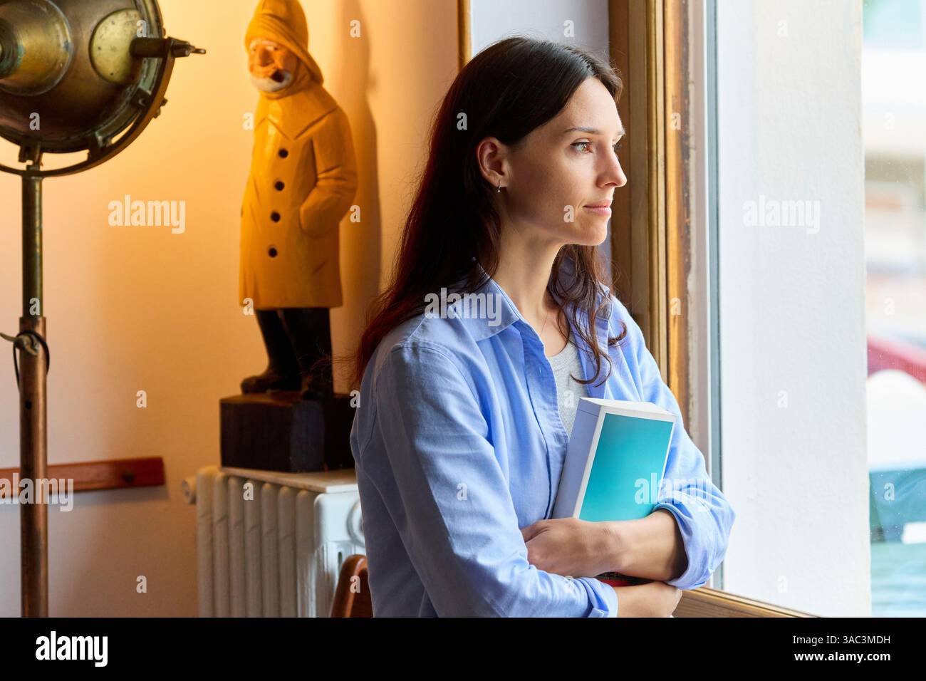 femme regardant par la fenêtre avec un livre dans les bras Banque D'Images