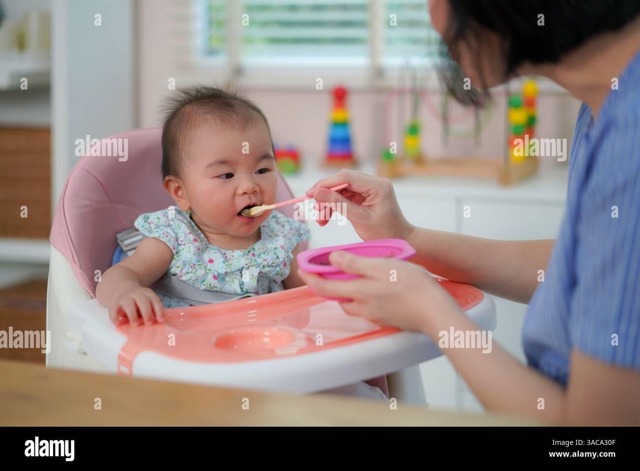 Un moment tendre capturant une mère nourrissant soigneusement son bébé à la cuillère tandis que le petit est assis dans une chaise haute rose. Banque D'Images