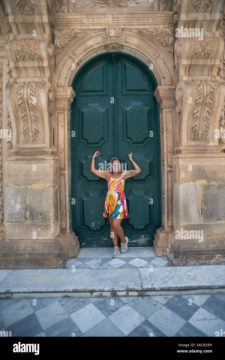Belle femme vêtue d'une robe blanche et de lunettes de soleil, souriante et heureuse, voyageant, appuyée contre une grande porte d'une église. Amérique du Sud. Banque D'Images