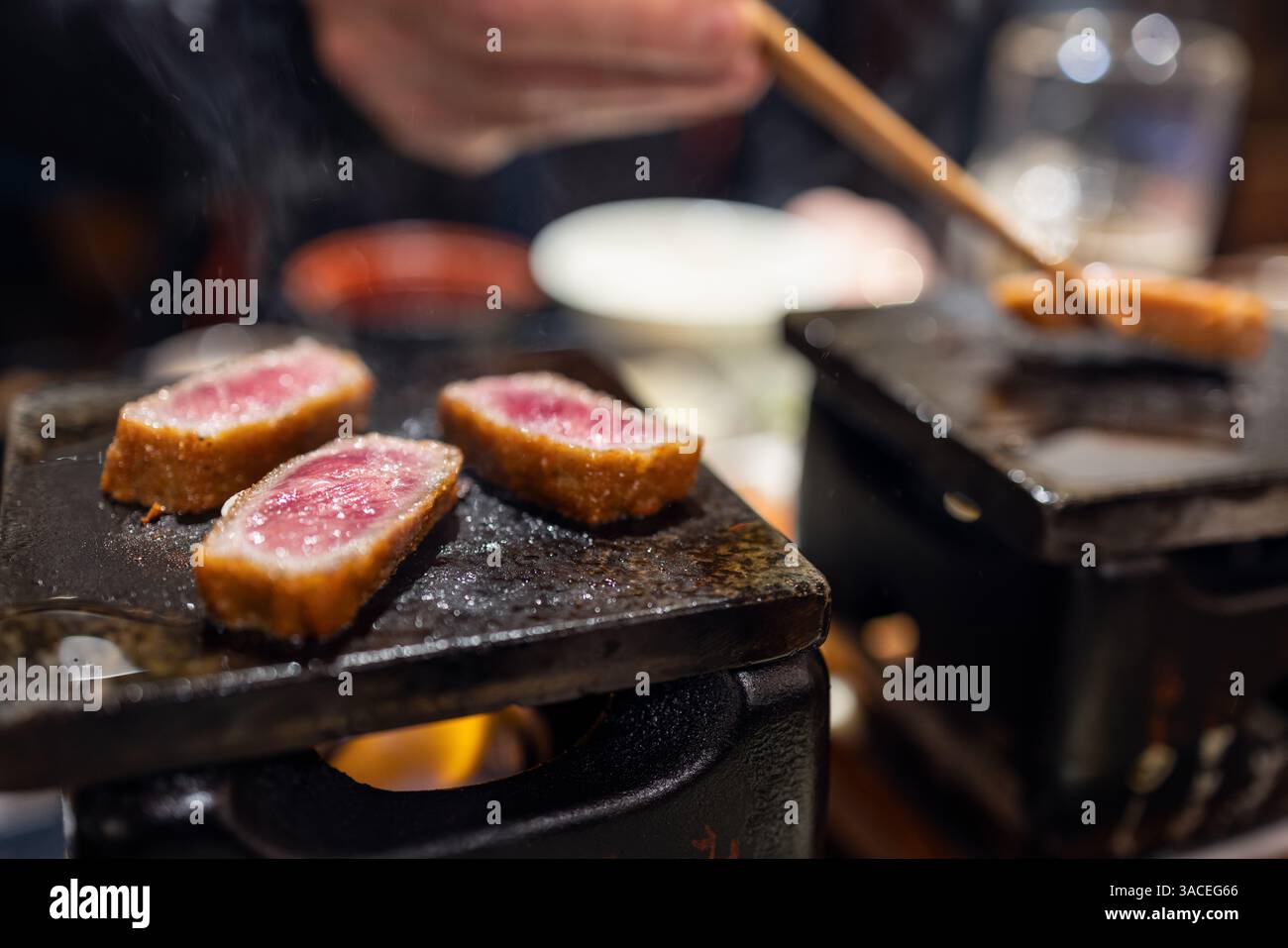 Trois morceaux de bœuf gyukatsu saisissant sur un gril en pierre de table avec la vapeur levant et une main utilisant des baguettes pour tourner une tranche sur le feu Banque D'Images
