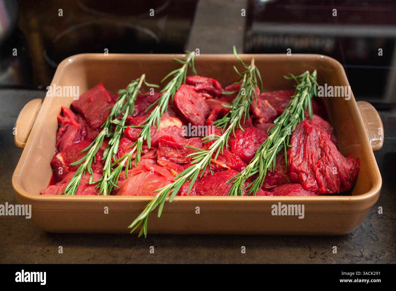 Morceaux de bœuf frais crus préparés dans un plat en céramique, garnis de brins de romarin, prêts à rôtir ou à cuire lentement. Banque D'Images