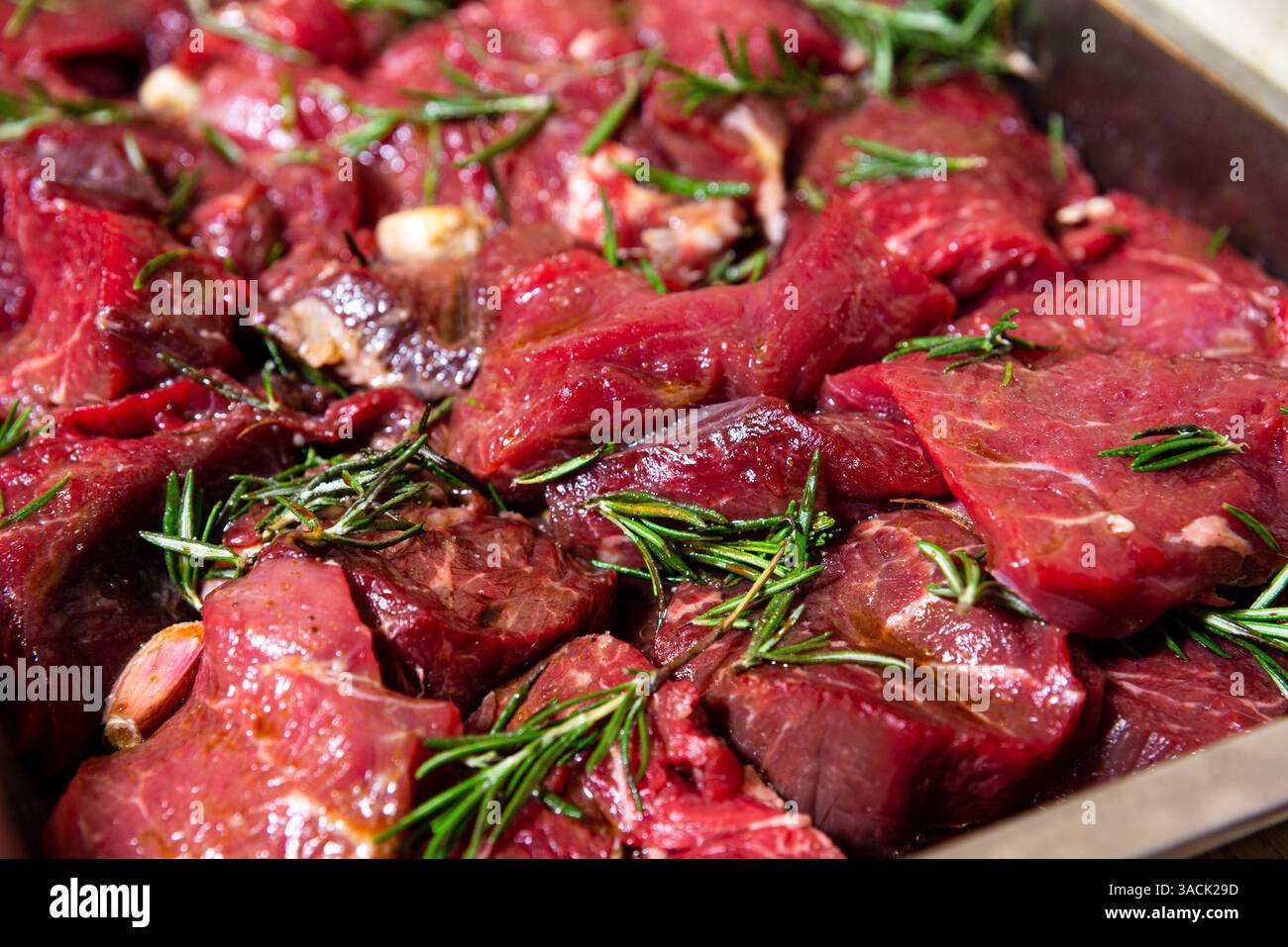 Gros plan de morceaux de boeuf frais crus dans un plateau, marinés avec de l'huile d'olive, de l'ail et du romarin pour la cuisson ou le grillage. Banque D'Images