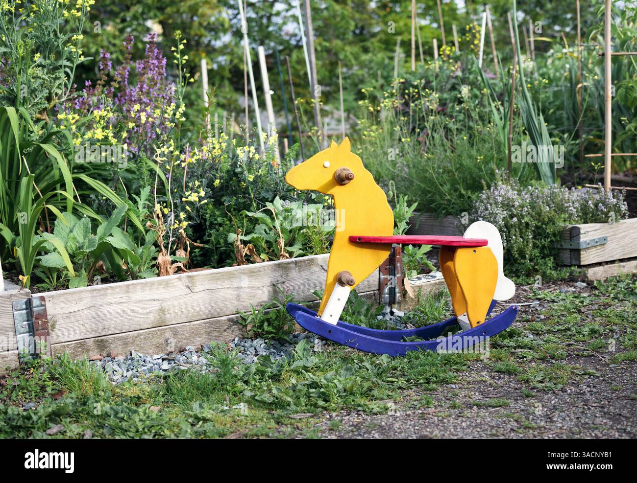 Cheval à bascule en bois dans le jardin. Les enfants vintage jouent à cheval devant des lits de jardin envahis par la végétation. Concept pour l'éducation à la nature, l'école immersive ou l'enfance Banque D'Images
