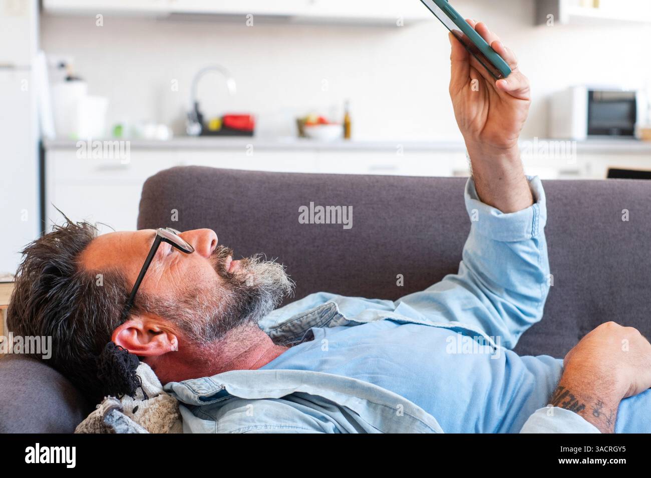 Homme allongé sur le canapé à la maison dans une position détendue, utilisant son téléphone pour envoyer des messages à des amis, faisant défiler le contenu multimédia divertissant et des vidéos tout en profitant de son temps libre Banque D'Images Homme allongé sur le canapé à la maison dans une position détendue, utilisant son téléphone pour envoyer des messages à des amis, faisant défiler le contenu multimédia divertissant et des vidéos tout en profitant de son temps libre Banque D'Images