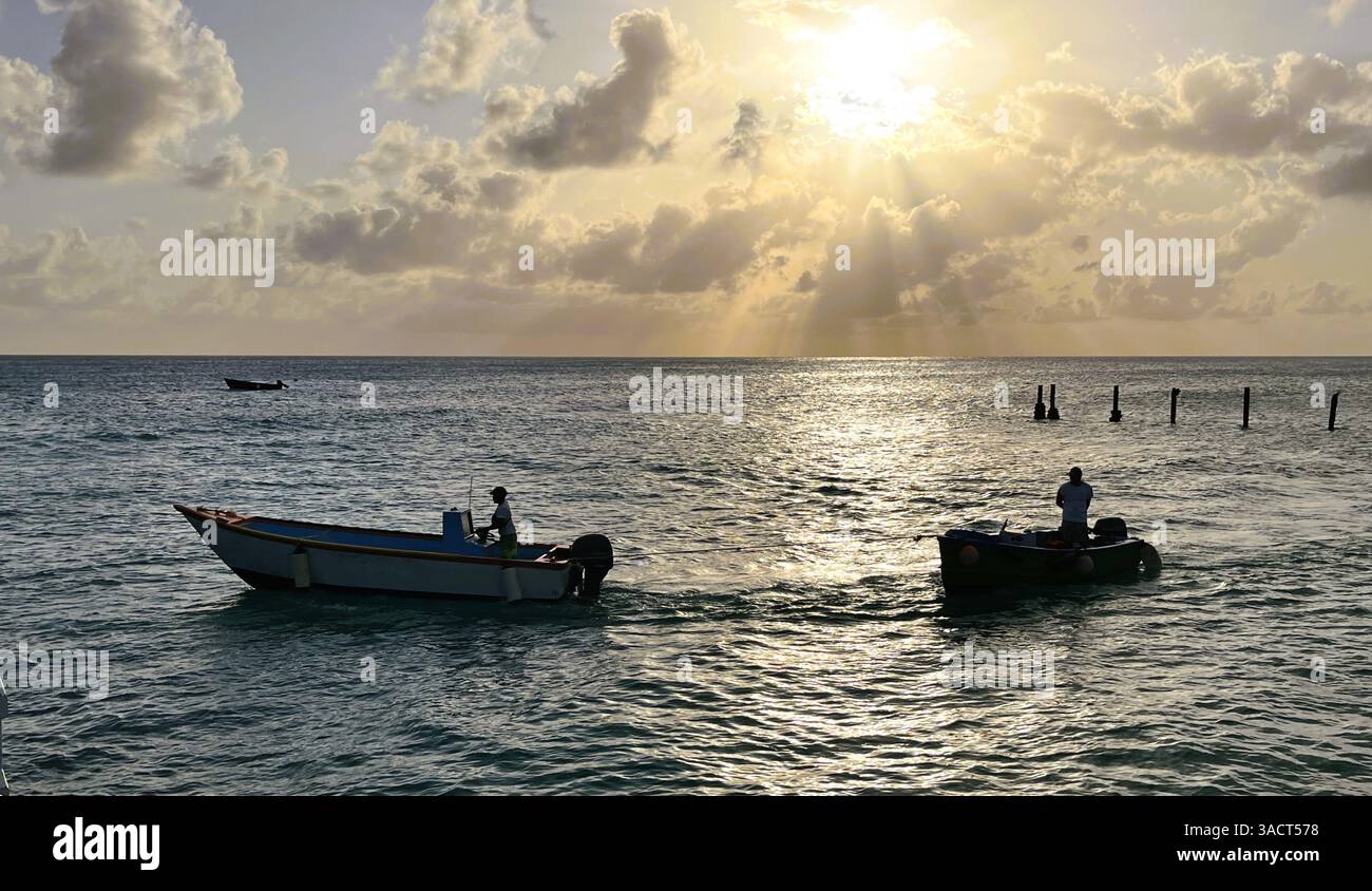 Un petit bateau remorque un autre bateau près d'Oistins, à la Barbade, alors que le soleil commence à se coucher derrière les nuages. Banque D'Images