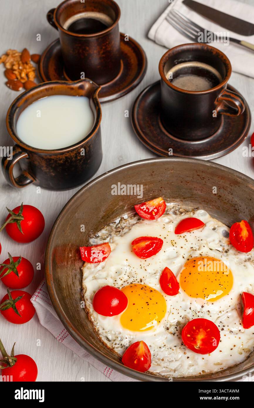 Deux œufs frits assaisonnés de poivre noir et décorés de tomates cerises sont servis dans une poêle pour un petit déjeuner sain et nutritif, accompagné de café, de lait et de noix Banque D'Images