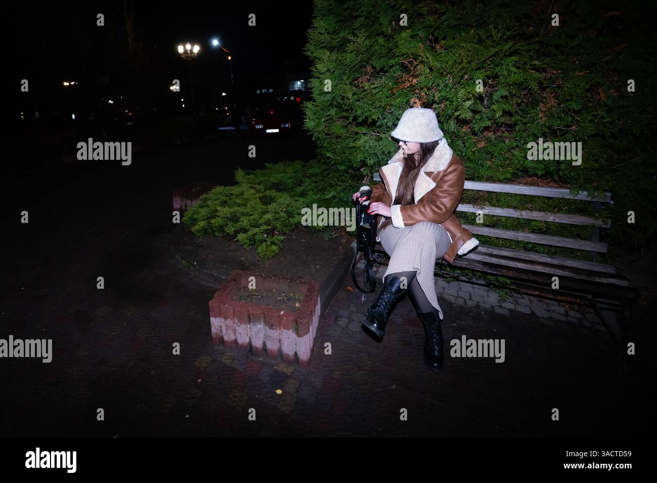 Sous la lueur des lampadaires, un individu à la mode est assis sur un banc de parc, habillé en tenue d'hiver, profitant d'un moment de calme entouré de gr luxuriants Banque D'Images