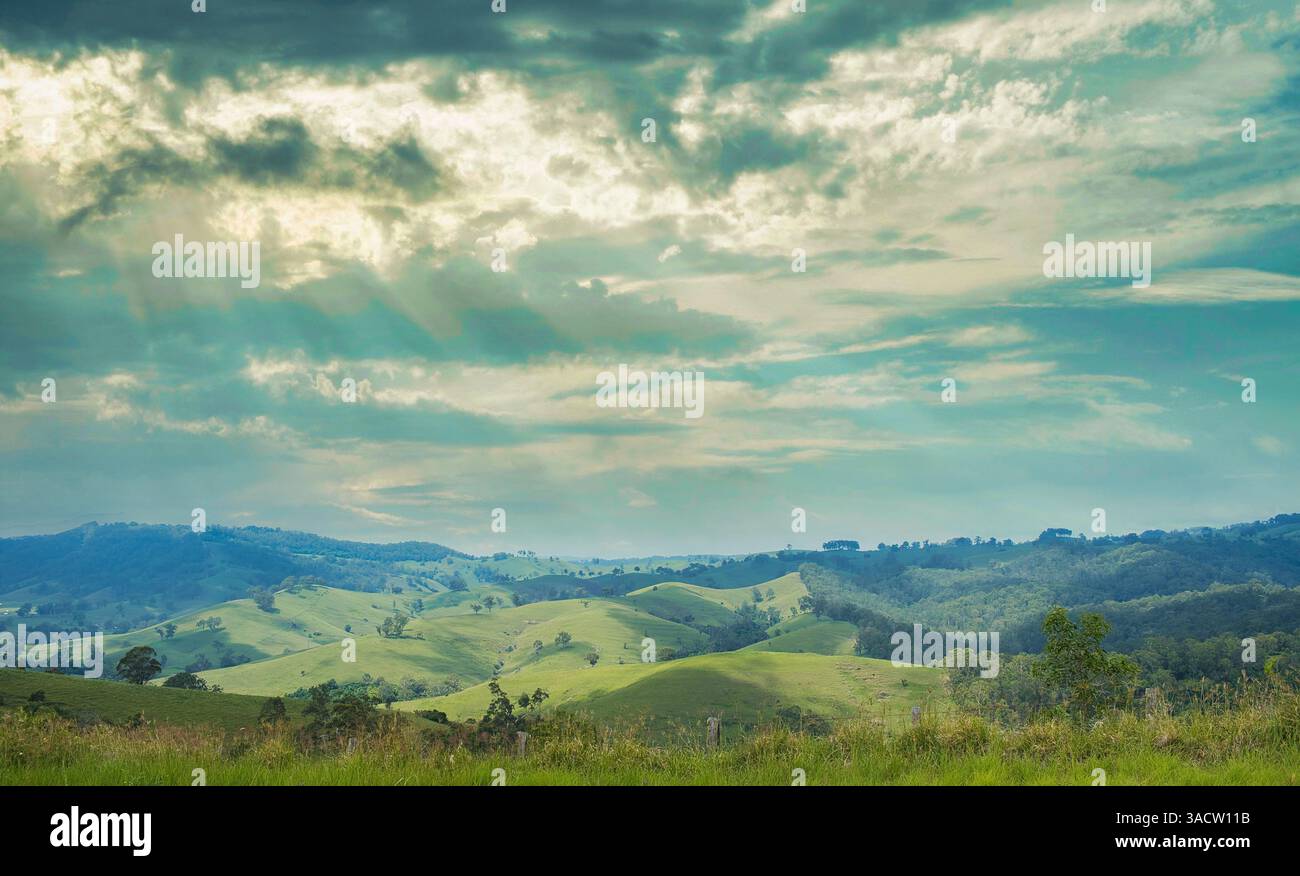 Paysage avec des collines, des prairies et des forêts aux couleurs vertes douces sous un ciel spectaculaire, avec des rayons de soleil qui brillent à travers les nuages. Banque D'Images