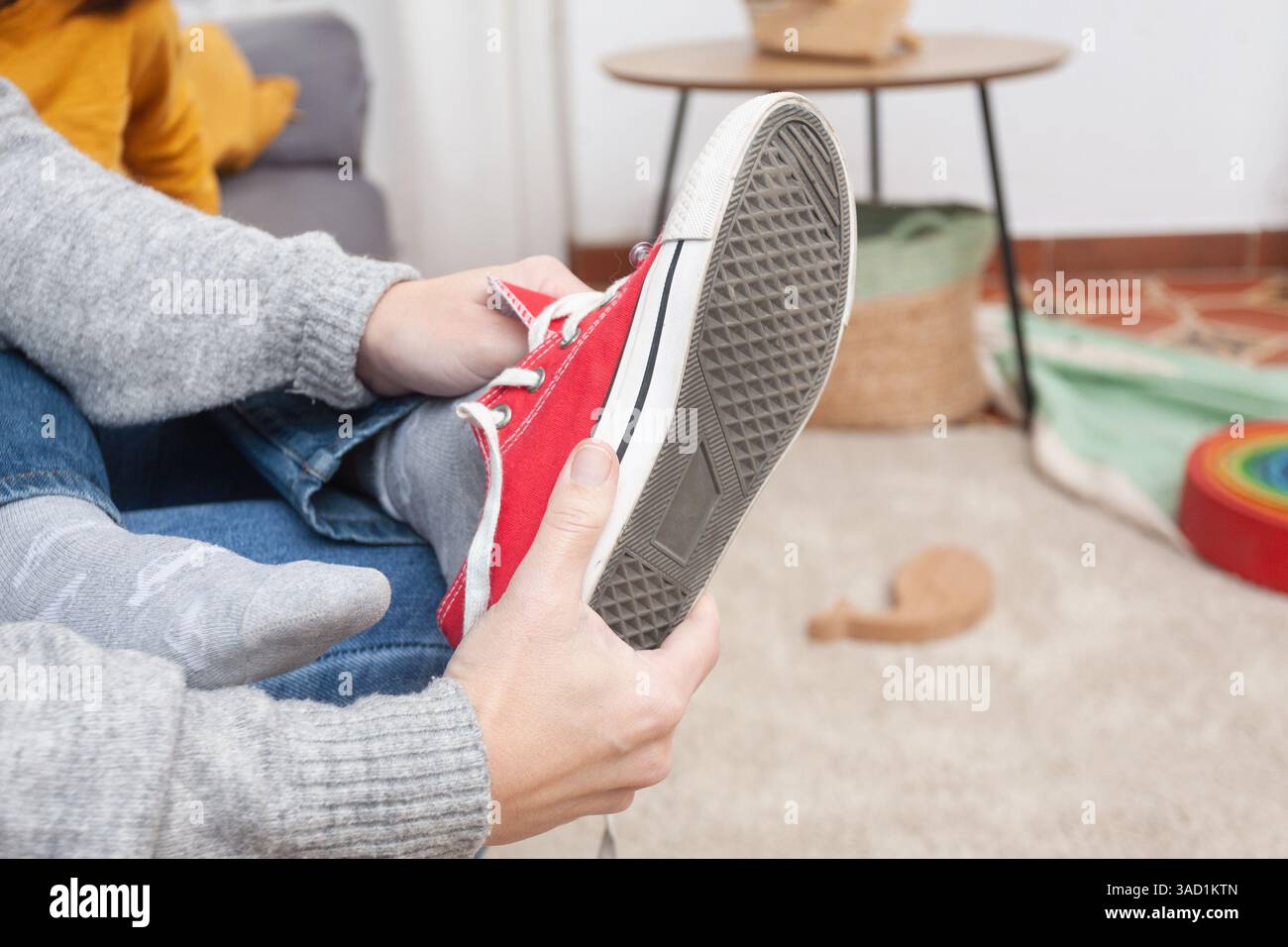 Un cliché détaillé du pied d’un enfant en tant que mère aide à enlever la sneaker rouge. Un geste subtil et émotionnel capturé dans un environnement familial chaleureux. Banque D'Images