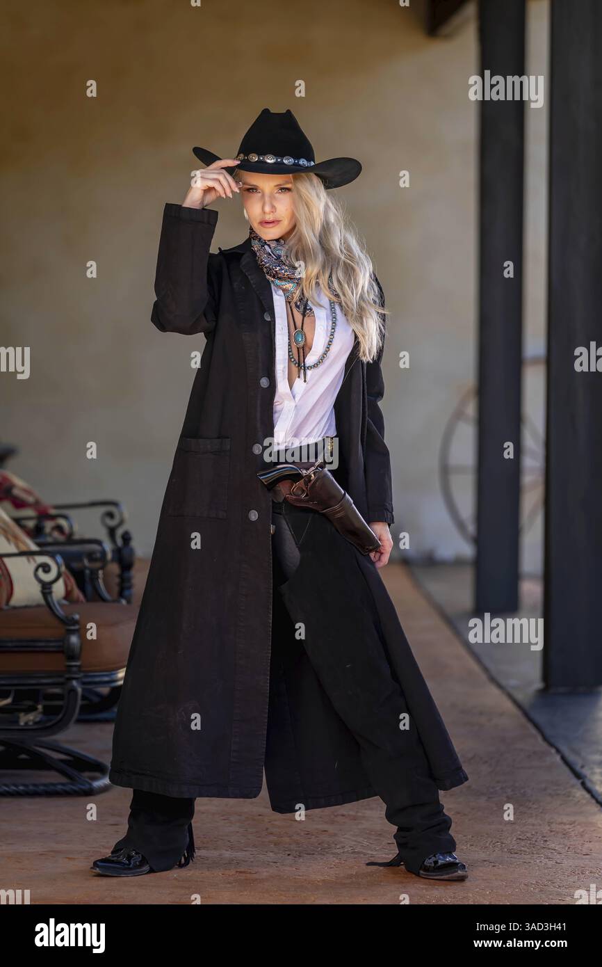 Une belle et confiante femme blonde dans une tenue de cow-boy classique se tient haute, posant avec un pistolet brillant sur sa hanche et un fusil reposant sur son shou Banque D'Images