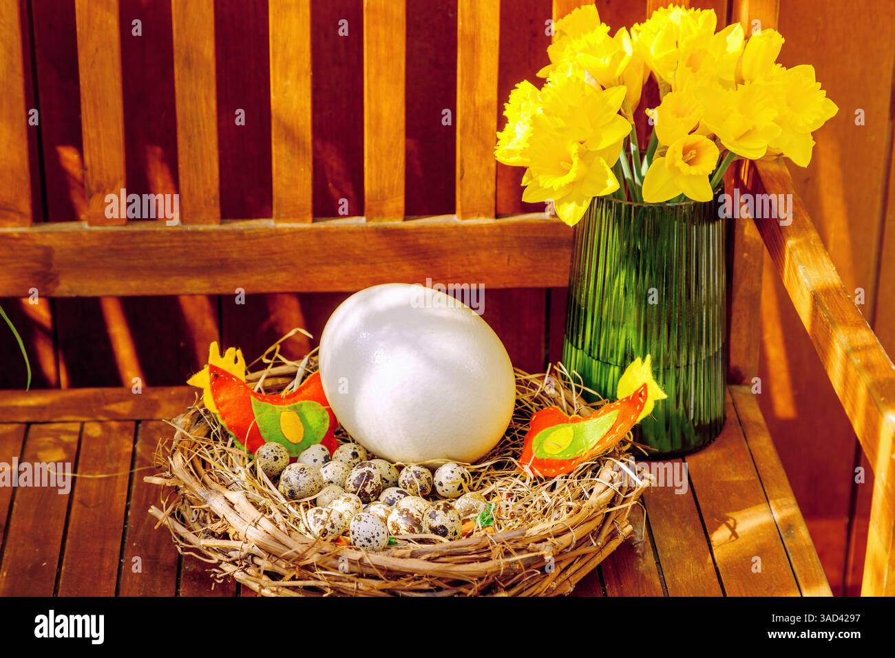Décorations de Pâques sur un banc en bois, nid de panier avec œufs de caille, œuf d'autruche et poulets en feutre faits maison, vase en verre avec jonquilles en fleurs Banque D'Images