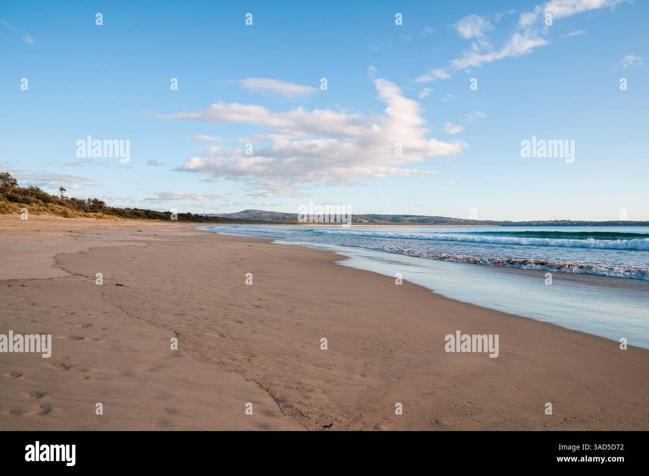 Plage de sable vide avec de douces vagues océaniques, collines lointaines, nuages doux et ciel bleu clair, littoral paisible, plage de Pambula, Australie Banque D'Images