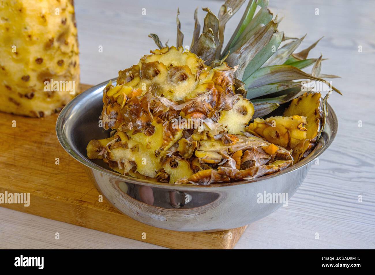 Un bol en acier inoxydable rempli de pelures d'ananas et la partie supérieure d'un ananas, placé sur une planche à découper en bois. Préparation de fruits tropicaux. Copier Banque D'Images