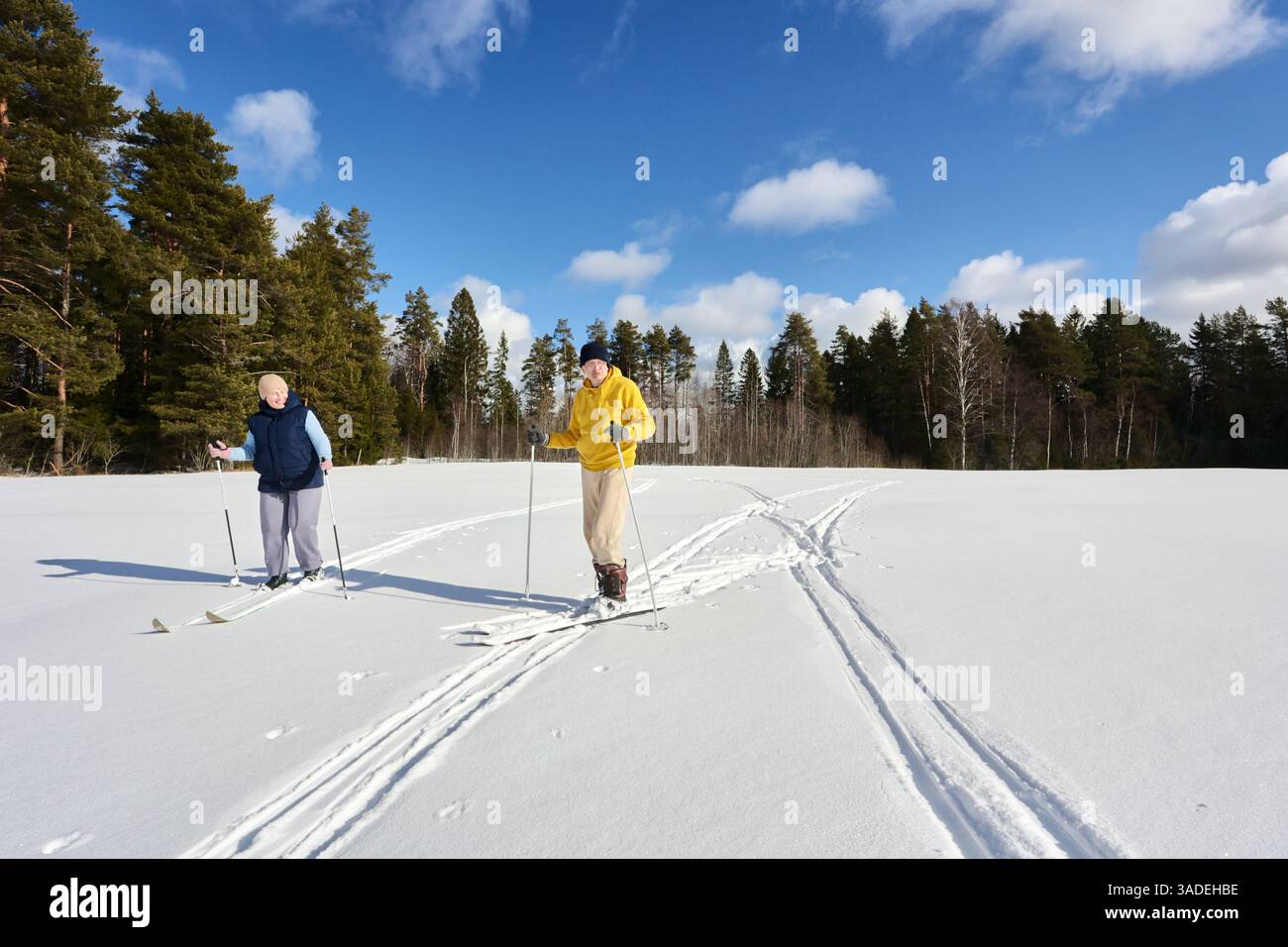 Les adultes plus âgés couple skis dans la neige, lui avec la veste jaune et elle traînant derrière, paire à peau claire profitant de l'exercice pendant la journée lumineuse, wint paisible Banque D'Images