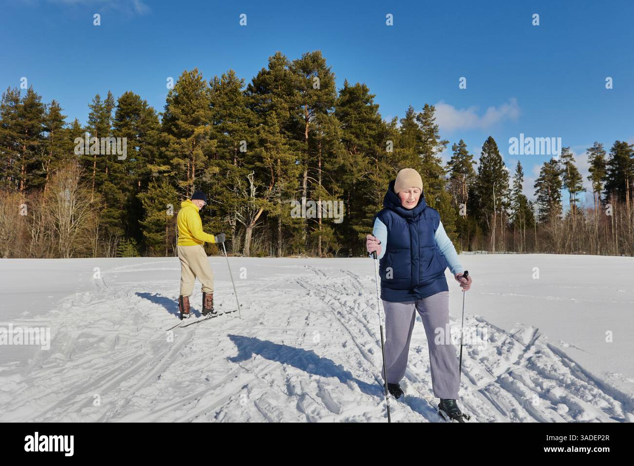 Mari mûr et femme dans les années 50 ski à travers le paysage enneigé, la femme prend la tête tandis que l'homme suit derrière, embrassant l'activité hivernale en plein air tog Banque D'Images