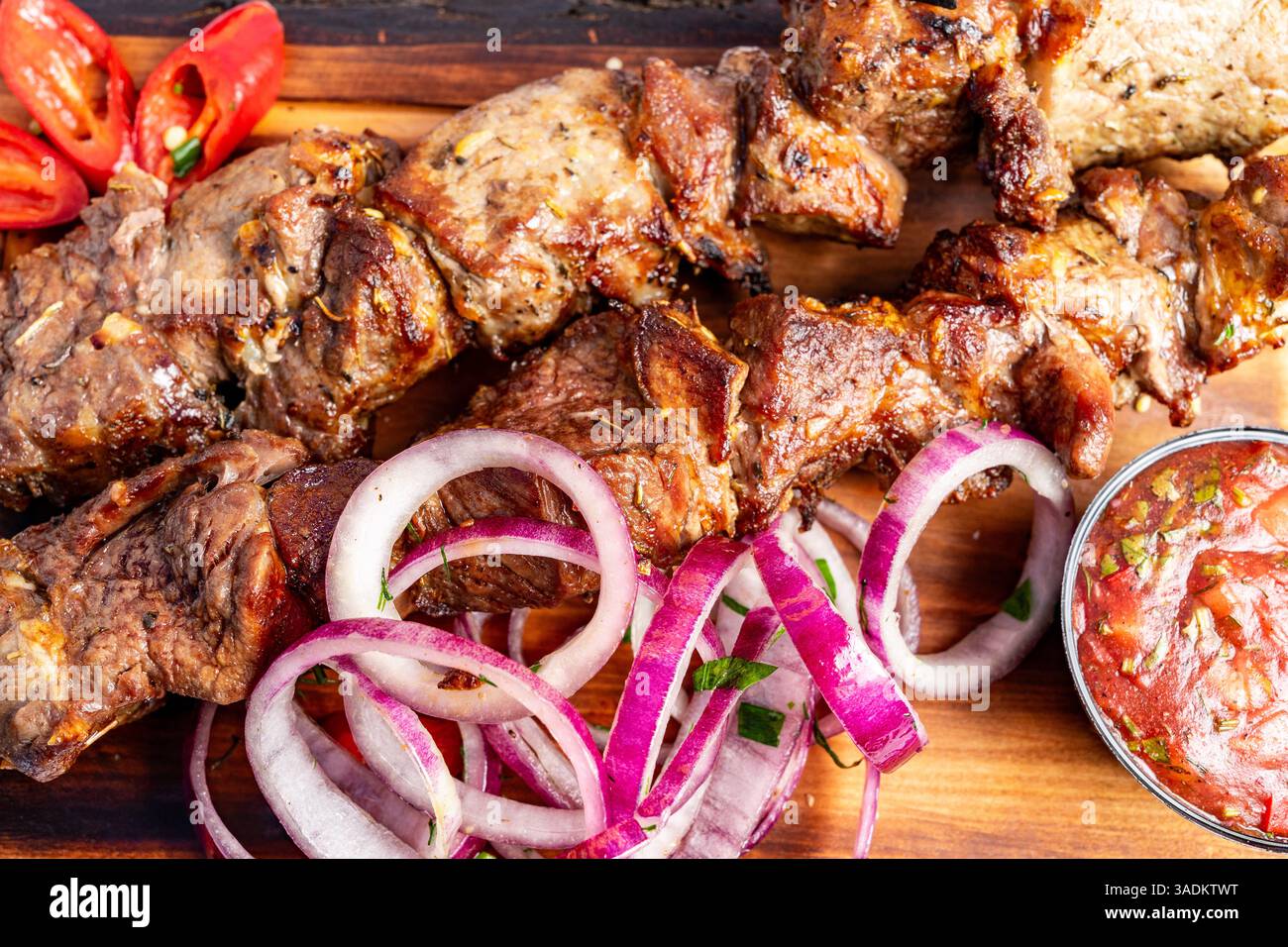 De succulentes brochettes de viande grillée sont servies avec des oignons rouges éclatants, des piments frais et une sauce salée. La présentation sur une planche de bois Banque D'Images