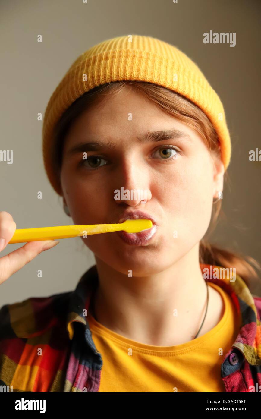 Une jeune femme hipster dans un chapeau jaune, se brossant les dents en gros plan. Son expression originale et son look moderne rendent le moment à la fois frais et stable Banque D'Images