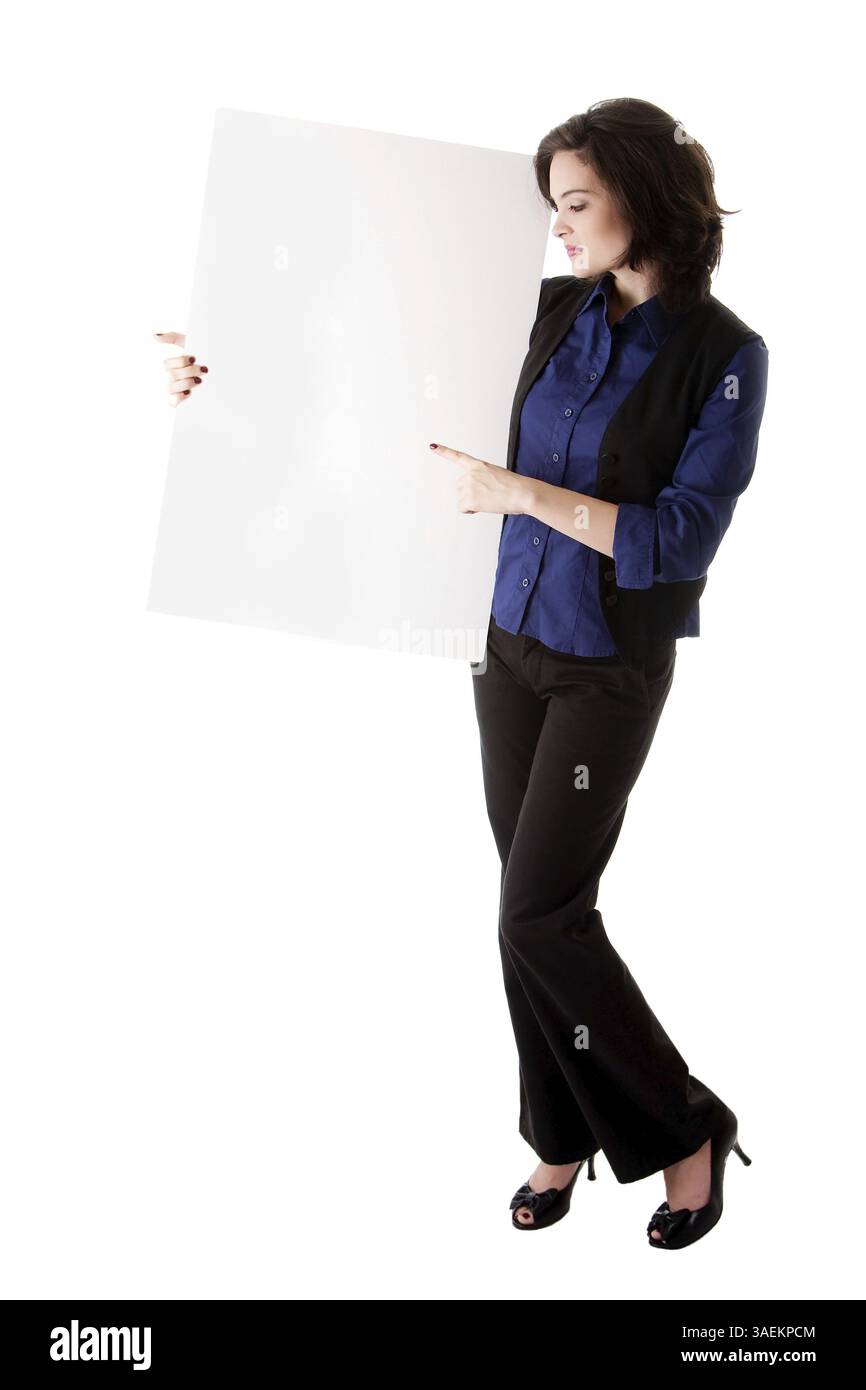 Happy smiling beautiful young caucasian brunette étudiant femme debout à côté et la tenue d'un conseil blanc vide et pointant vers elle, isolée Banque D'Images