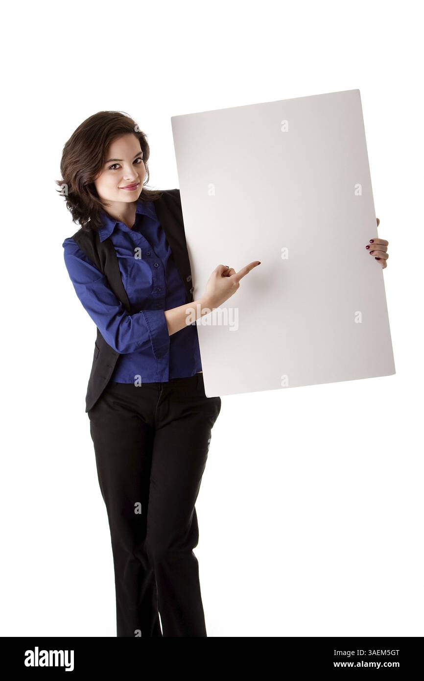 Happy smiling beautiful young caucasian brunette étudiant femme debout à côté et la tenue d'un conseil blanc vide et pointant vers elle, isolée Banque D'Images