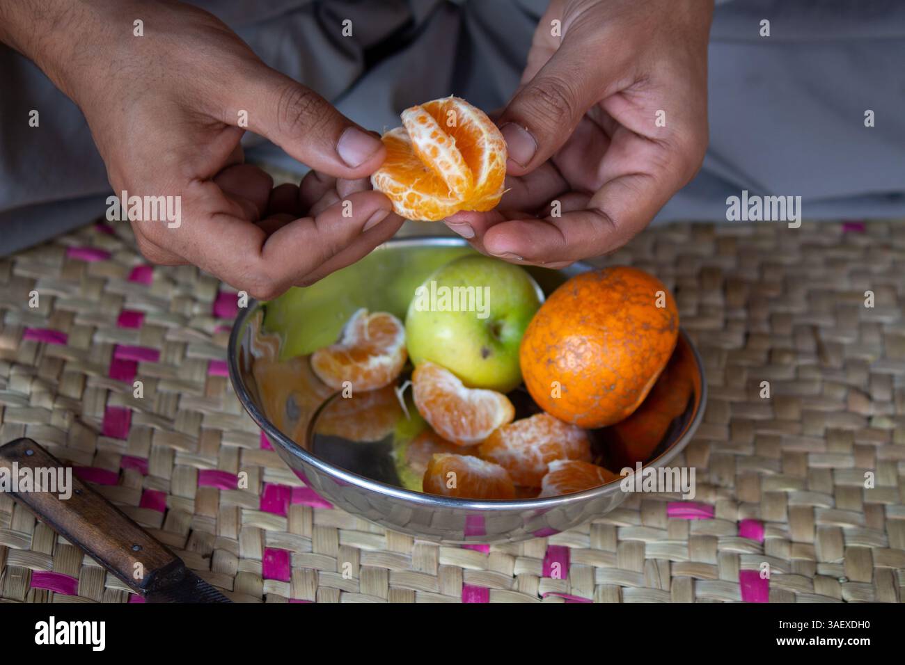 Une personne épluchant une orange fraîche avec les mains, bol en acier inoxydable avec des pommes et des oranges sur un tapis tissé, représentant un mode de vie rural. Banque D'Images