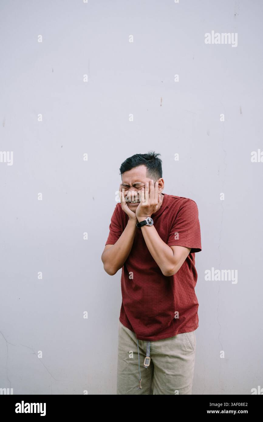 Homme asiatique stressé avec douleur insupportable mal de dents serrant son visage avec les deux mains contre un mur plat en plein air Banque D'Images