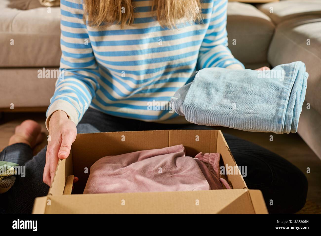 Femme emballe boîte de don en carton avec des vêtements dans le salon. Personne préparant des vêtements usagés pour le recyclage ou l'occasion. Concept de charité, d'aide sociale et de bénévolat Banque D'Images