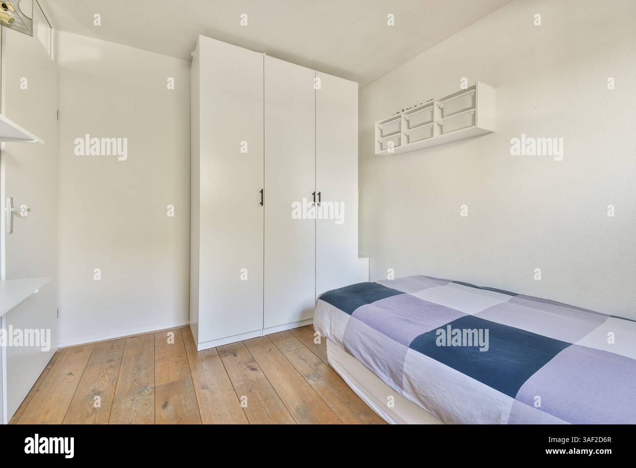 Une chambre lumineuse et minimaliste avec une armoire blanche contre un mur clair et un couvre-lit à carreaux, mettant l'accent sur la simplicité et le calme. Banque D'Images