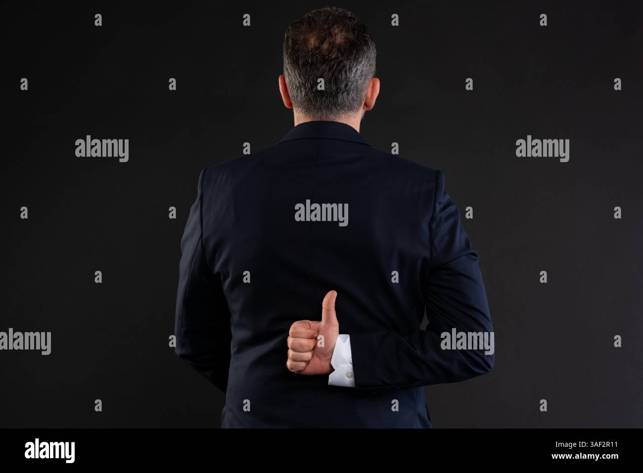 Homme en costume vu de derrière faisant un geste secret du pouce vers le haut, symbolisant l'approbation cachée, l'ironie ou le double sens dans un contexte d'affaires. Banque D'Images