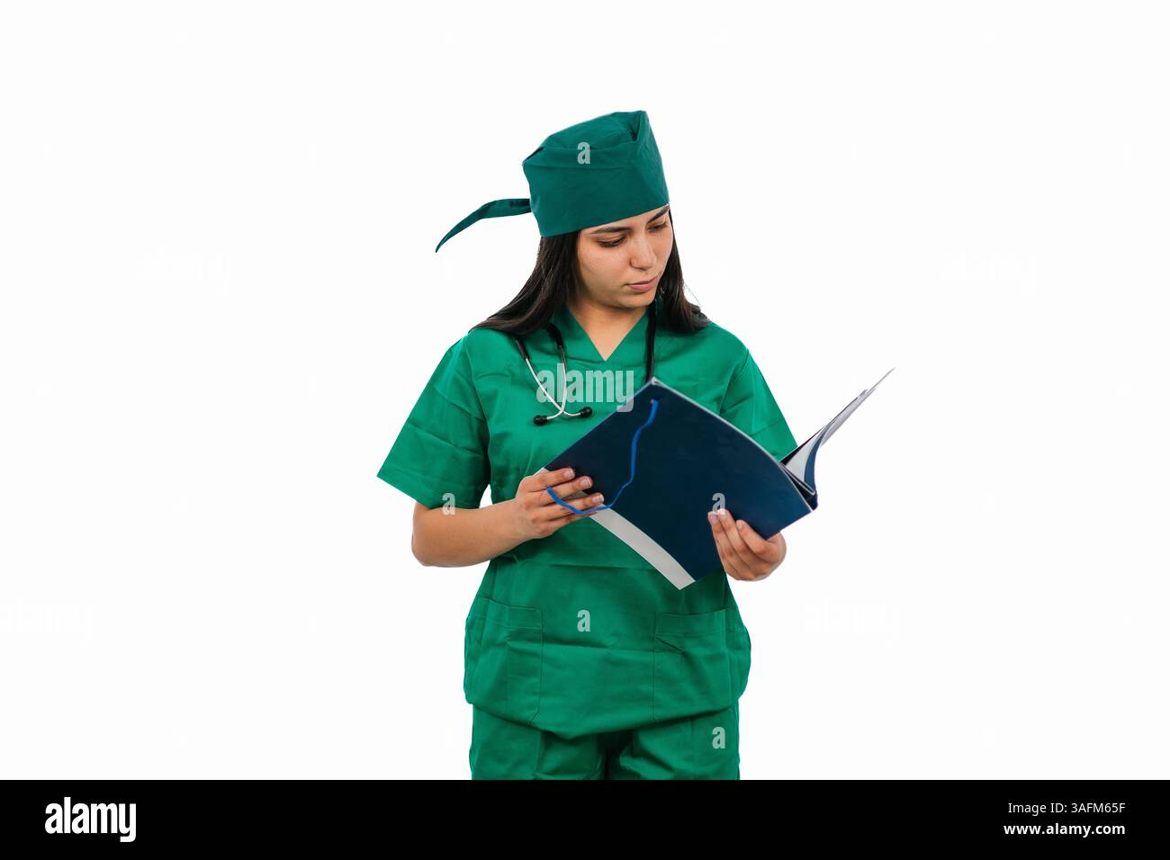 Une jeune femme chirurgienne portant des gommages verts et un stéthoscope lit attentivement un livre de dossier médical, isolé sur un fond blanc, fournissant un clea Banque D'Images