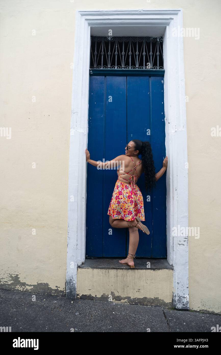Belle jeune femme caucasienne avec des tresses, des lunettes de soleil et une robe colorée debout par une porte bleue d'une vieille maison. Personne confiante. Brésil Banque D'Images