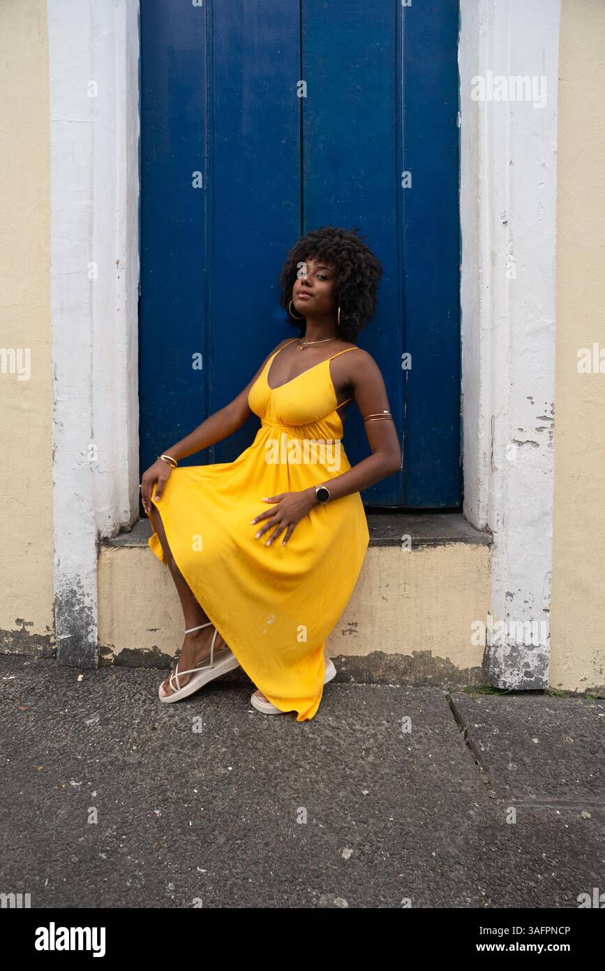 Belle jeune femme aux cheveux afro, vêtue d'une robe jaune, assise près d'une porte bleue d'une vieille maison. Personne confiante. Brésil Banque D'Images