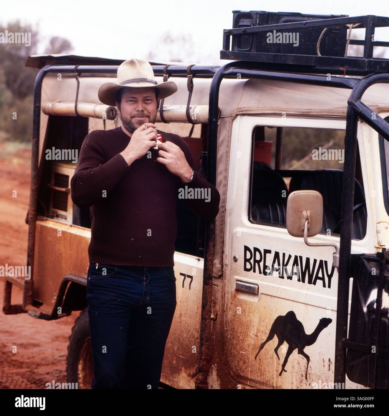 Mann im australischen Outback, der entspannt vor einem staubigen Geländewagen mit der Aufschrift 'Breakaway' steht. Er trägt einen typischen Cowboyhut und einen dunklen Pullover, während er genüsslich eine Zigarette raucht. Der Offroad-wagen mit Staub- und Schlammspritzern deutet darauf Hin, dass er auf einer langen Expedition oder Offroad-Tour unterwegs ist. Die Szenerie vermittelt ein Gefühl von Abenteuer, Freiheit und Wildnis, um 1985. Banque D'Images