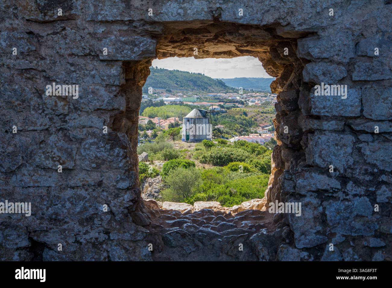 Vieux mur de pierre encadrant une vue panoramique sur un moulin à vent traditionnel et un paysage luxuriant à Óbidos, Portugal. Un aperçu rustique du charme rural européen. Banque D'Images