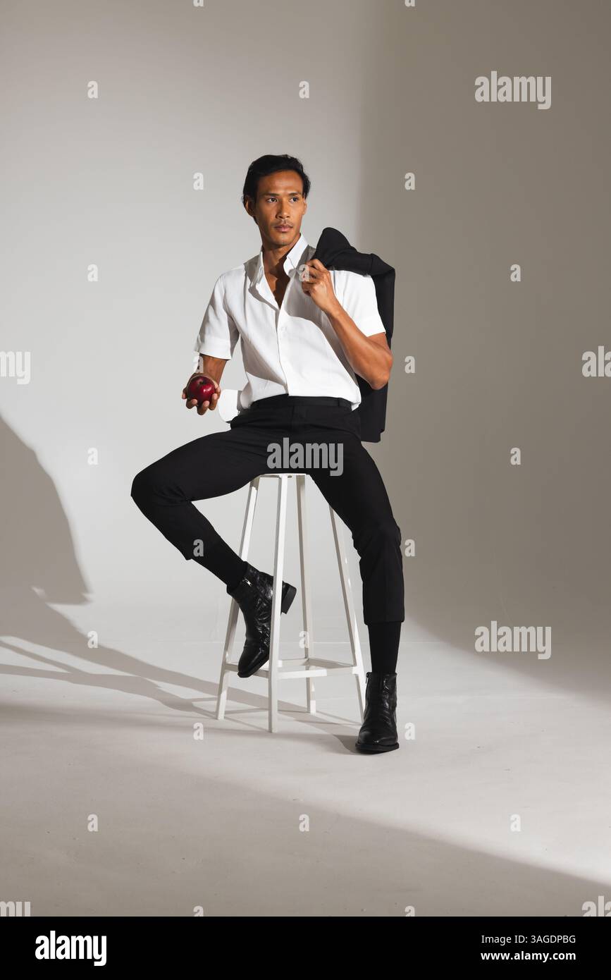 Homme confiant dans une tenue élégante tenant la pomme, assis sur le tabouret dans le studio. Mode, confiance, style de vie, vêtements, portrait, masculin Banque D'Images