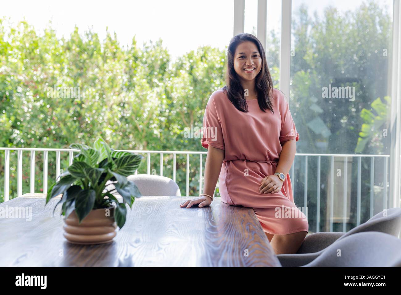 Femme souriante en robe rose assise sur la table près de la grande fenêtre à la maison. Style de vie, mode, féminin, élégance, loisirs, relaxation Banque D'Images