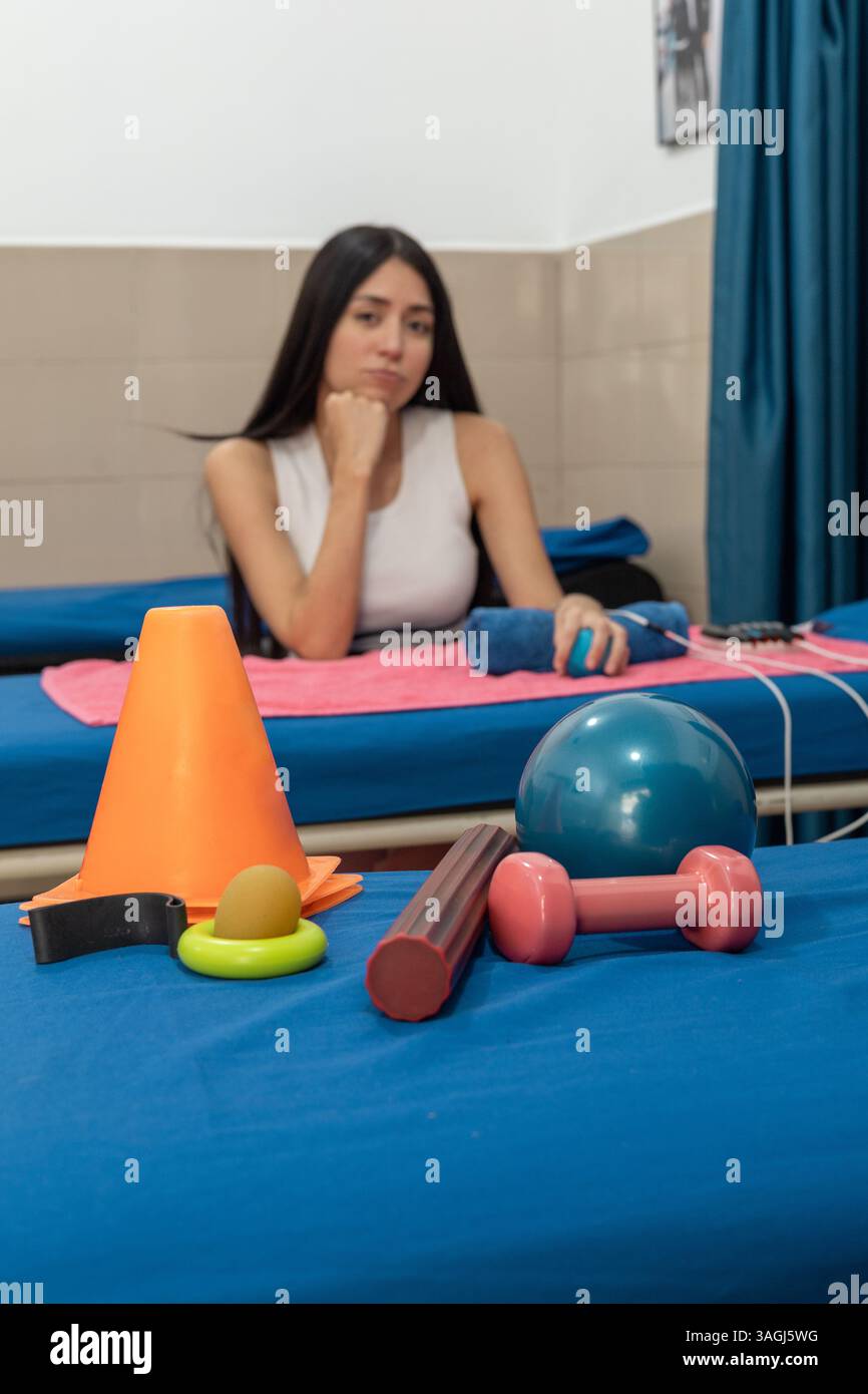 Une femme reçoit de l'électrothérapie sur son bras tandis que divers outils colorés de physiothérapie sont placés au premier plan. Banque D'Images