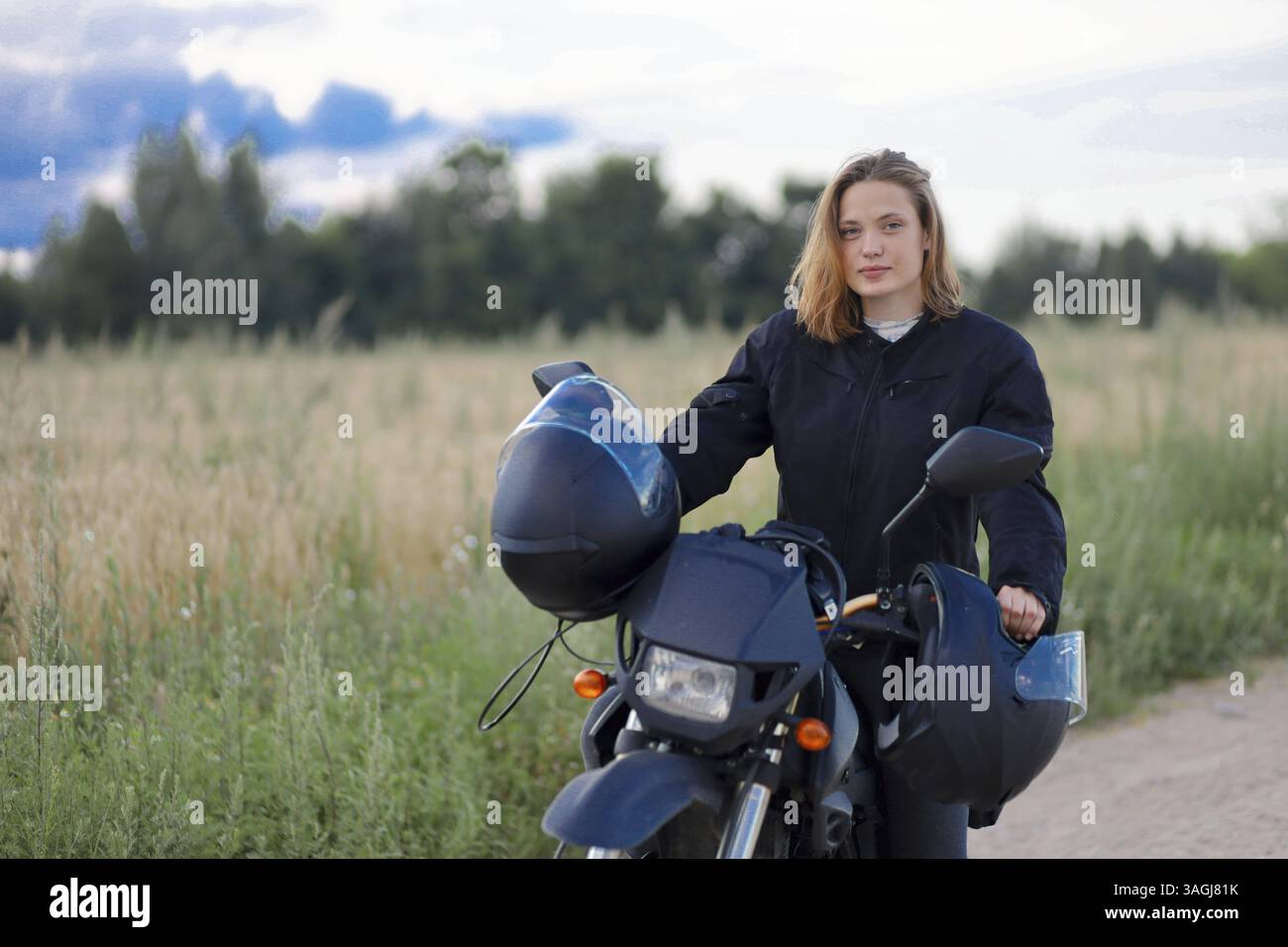 Une femme debout à côté d'une moto dans un paysage rural, portant une veste noire et tenant un casque Banque D'Images