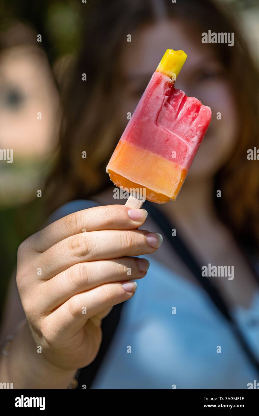 Main de filles tenant mordu fruit rouge jaune popsicle crème glacée sur le bâton en bois. Arôme fraise, cerise, orange, mangue. Journée d'été ensoleillée dans le parc. Banque D'Images