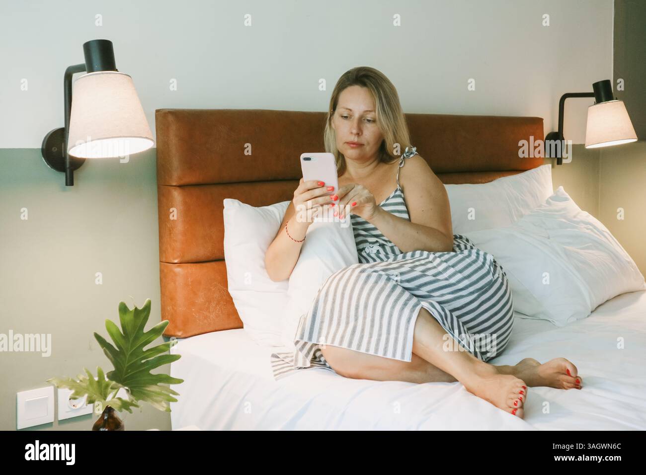 Belle femme blonde à l'aide d'un smartphone couché sur le lit dans la chambre Banque D'Images