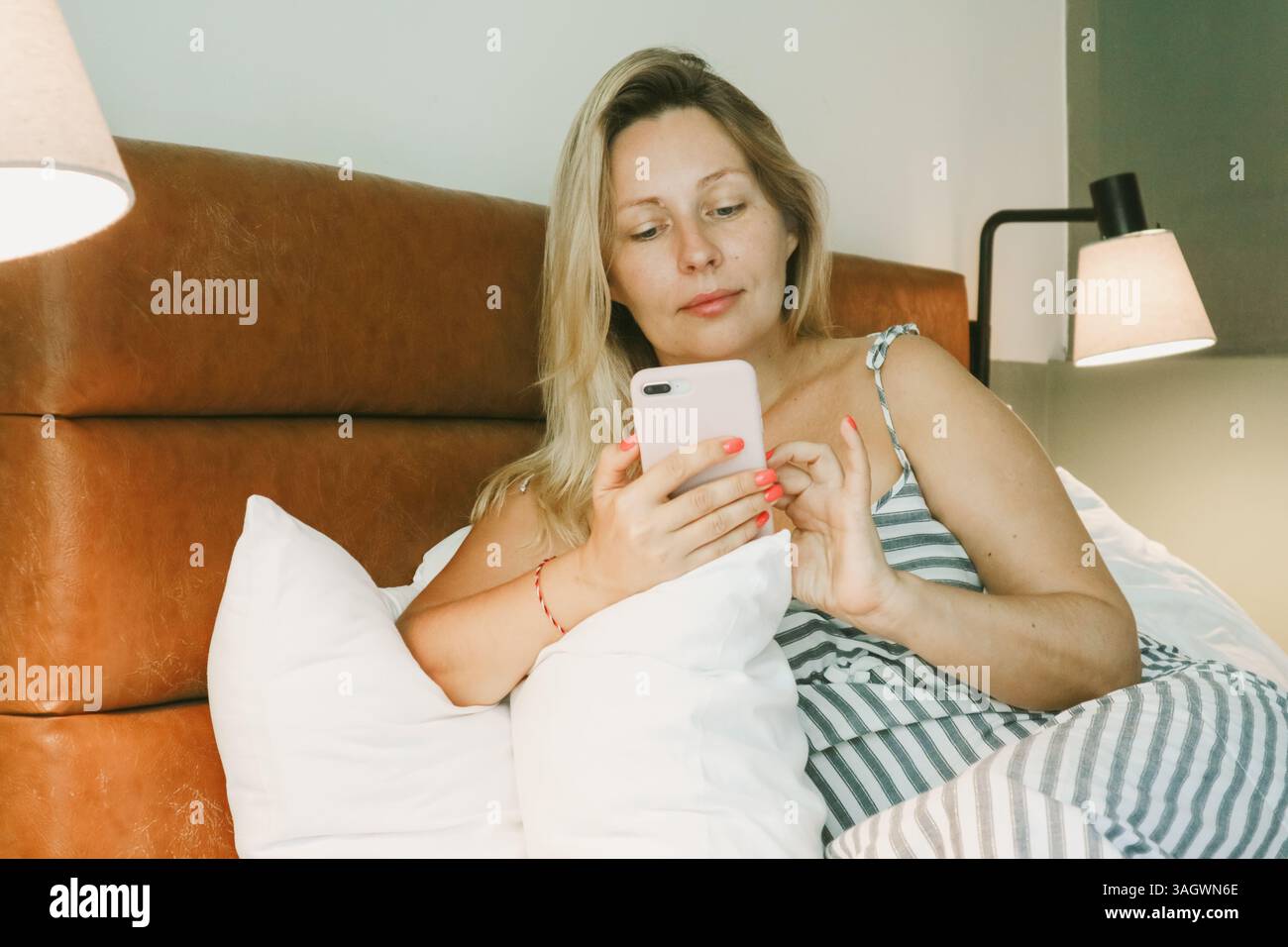Belle femme blonde à l'aide d'un smartphone couché sur le lit dans la chambre Banque D'Images