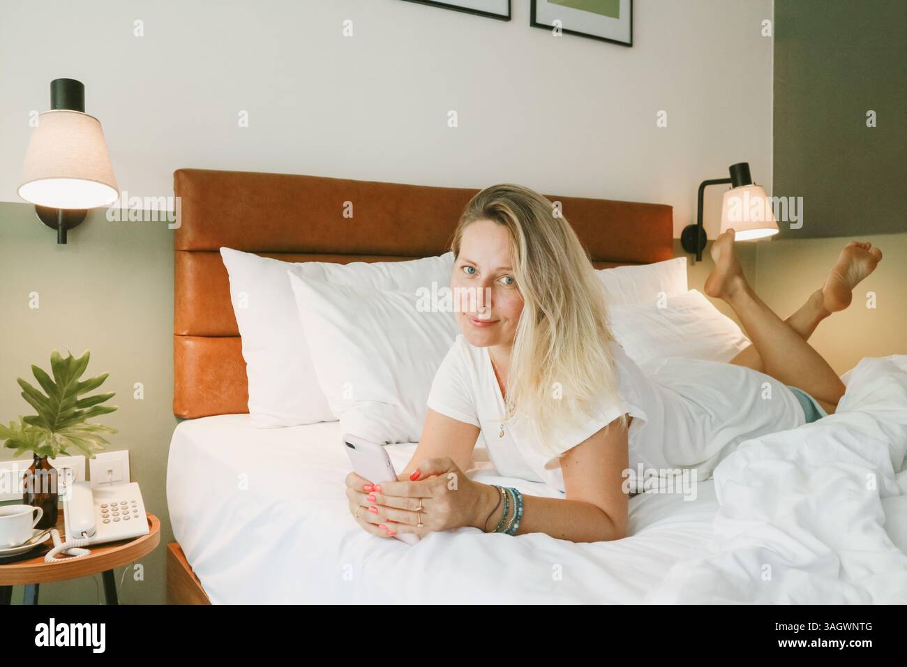 Belle femme blonde à l'aide d'un smartphone couché sur le lit dans la chambre Banque D'Images