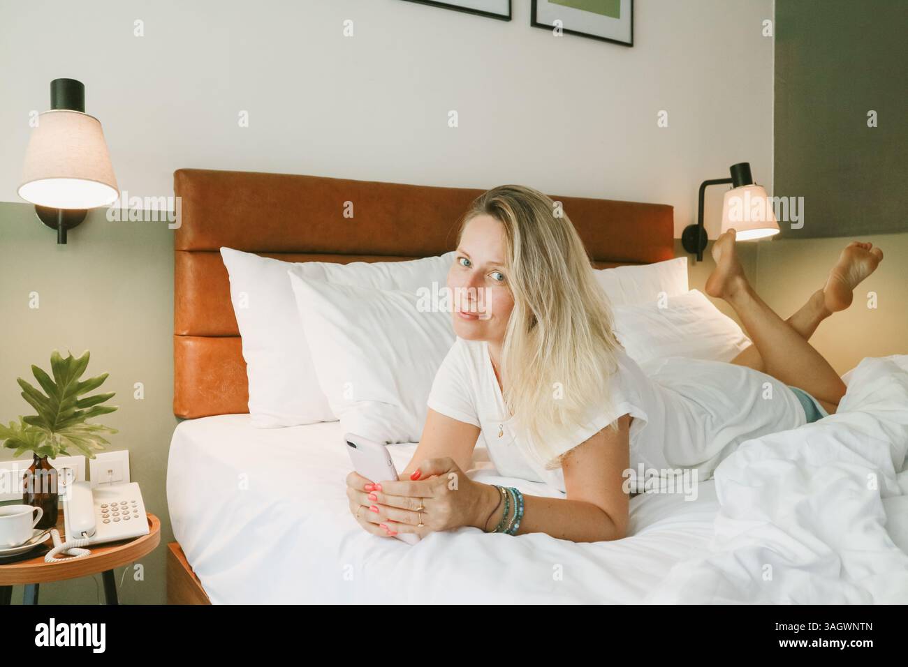 Belle femme blonde à l'aide d'un smartphone couché sur le lit dans la chambre Banque D'Images