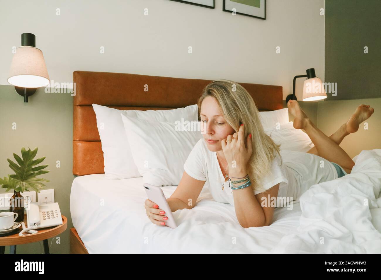 Belle femme blonde à l'aide d'un smartphone couché sur le lit dans la chambre Banque D'Images