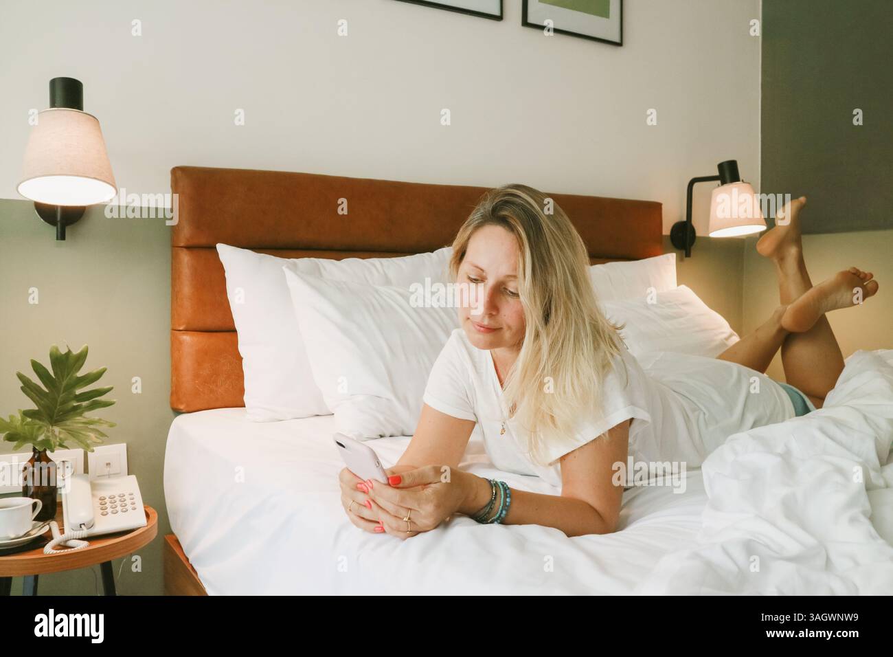 Belle femme blonde à l'aide d'un smartphone couché sur le lit dans la chambre Banque D'Images