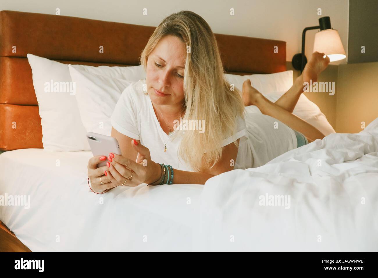 Belle femme blonde à l'aide d'un smartphone couché sur le lit dans la chambre Banque D'Images