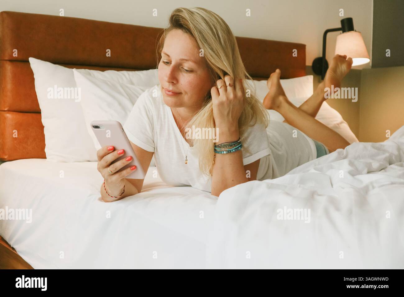 Belle femme blonde à l'aide d'un smartphone couché sur le lit dans la chambre Banque D'Images