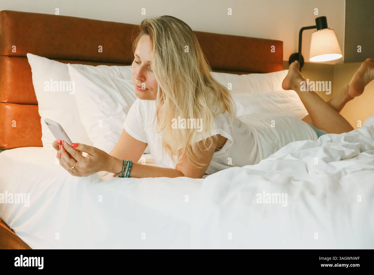 Belle femme blonde à l'aide d'un smartphone couché sur le lit dans la chambre Banque D'Images