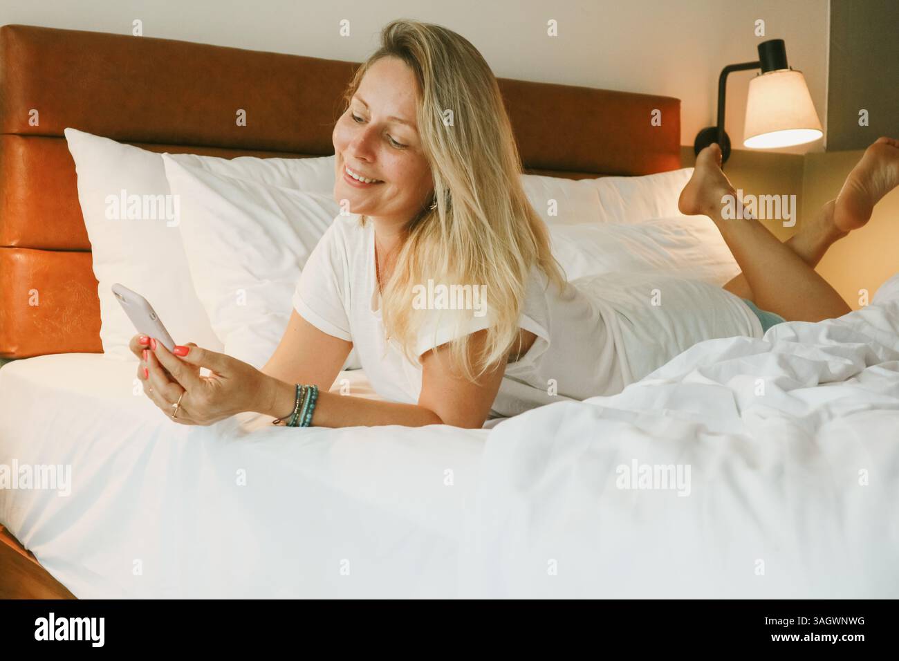 Belle femme blonde à l'aide d'un smartphone couché sur le lit dans la chambre Banque D'Images